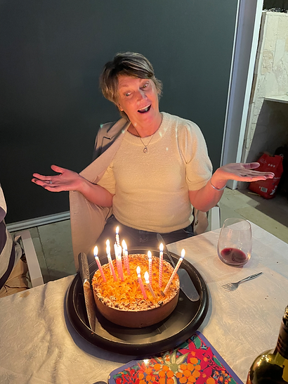 Michelle Martin di meja makan mengangkat bahu bermain -main di belakang kek hari jadi yang diliputi dalam lilin yang menyala; Pencahayaan dalaman hangat, kaca wain dan botol berdekatan.